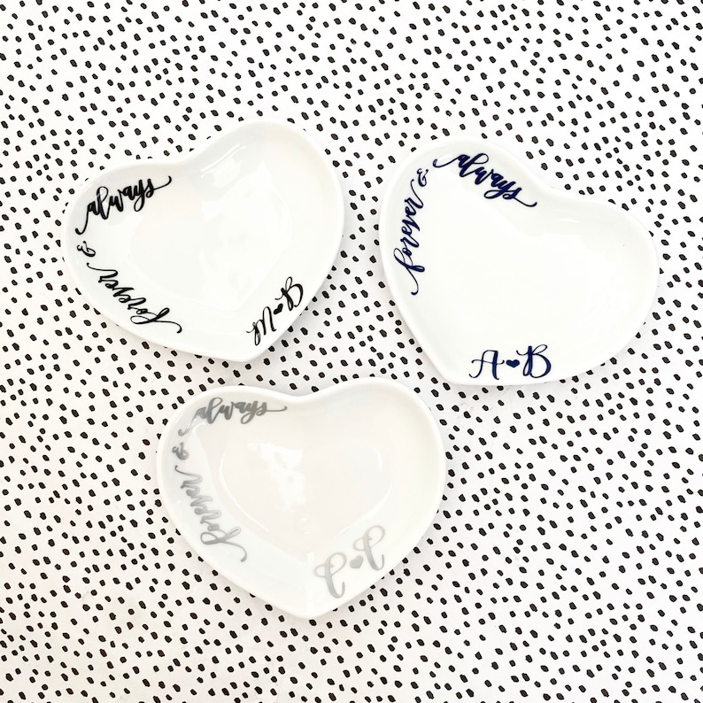 May include: Three white heart-shaped ceramic dishes with black and silver lettering. The dishes are decorated with the words "always & forever" and "always & married" and the initials "A-B", "M-B", and "C-F".