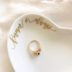May include: A white heart-shaped dish with gold lettering that reads "Forever & Always". A gold ring with a black stone and small diamonds is sitting in the dish.
