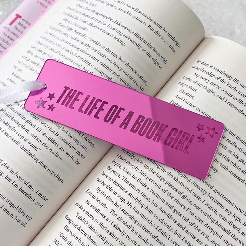 May include: A bright pink bookmark with the text "THE LIFE OF A BOOK GIRL" in a bold font, along with star accents. The bookmark is rectangular and lies on the open pages of a book, with a white ribbon attached.