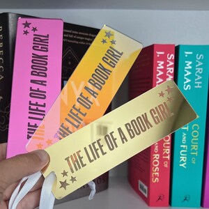 May include: Three rectangular bookmarks with the text "THE LIFE OF A BOOK GIRL" and star accents. The bookmarks are pink, orange, and gold, with white ribbon tassels. Books with colorful spines are in the background.