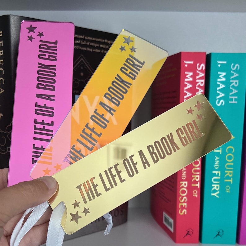 May include: Three rectangular bookmarks with the text "THE LIFE OF A BOOK GIRL" and star accents. The bookmarks are pink, orange, and gold, with white ribbon tassels. Books with colorful spines are in the background.