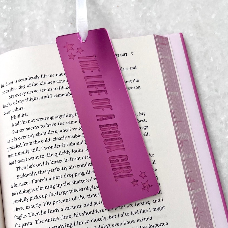 May include: A vibrant magenta bookmark with the text "THE LIFE OF A BOOK GIRL" and star accents. The bookmark is made of a reflective material and has a white ribbon attached. It rests on the pages of a book.