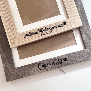 May include: Two rectangular picture frames with white matting. The top frame is light wood with black text reading Anderson Mobile Grooming Est. 2024 and a paw print. The bottom frame is dark gray wood with black text reading Cabo & Cali and a paw print.