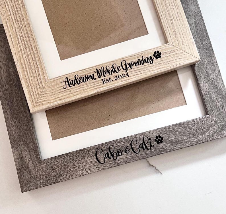 May include: Two rectangular picture frames with white matting. The top frame is light wood with black text reading Anderson Mobile Grooming Est. 2024 and a paw print. The bottom frame is dark gray wood with black text reading Cabo & Cali and a paw print.