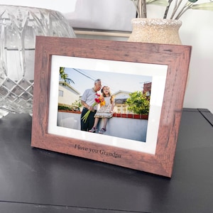 May include: A brown wooden picture frame with a white mat and a photo of an older man and a young girl smiling. The photo is of the two standing on a brick wall with flowers in the man's hand. The frame has the words "I love you Grandpa" engraved on the bottom.