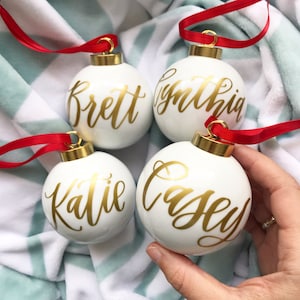 May include: Four white Christmas ornaments with gold lettering. The ornaments are decorated with the names "Brett", "Cynthia", "Katie", and "Casey".