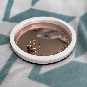 May include: A white ring dish with a rose gold interior and the text "story (Paige's version)" inscribed on the side. A gold ring with black stones is resting in the dish.