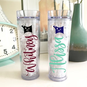 May include: Two clear plastic tumblers with a mortar and pestle design and the names "Whitney" and "Alyssa" written in cursive. The tumblers are filled with white crinkle paper.