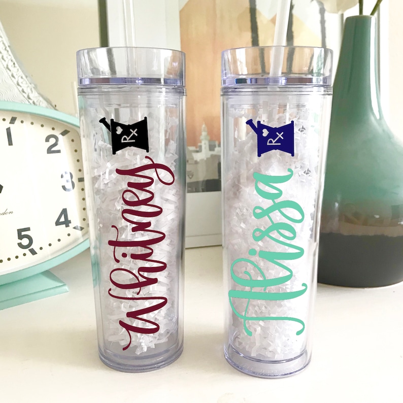 May include: Two clear plastic tumblers with a mortar and pestle design and the names "Whitney" and "Alyssa" written in cursive. The tumblers are filled with white crinkle paper.