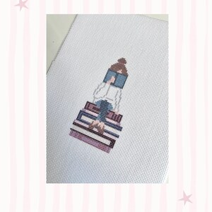 K&ouml;nnte beinhalten: Kreuzstich-Kunstwerk, das eine Person zeigt, die auf einem Stapel B&uuml;cher sitzt und liest. Die Figur ist in einem minimalistischen Stil mit einer Palette aus Blau-, Lila- und Braunt&ouml;nen dargestellt. Der Hintergrund ist eine wei&szlig;e Leinwand mit einem rosa-wei&szlig; gestreiften Rand.