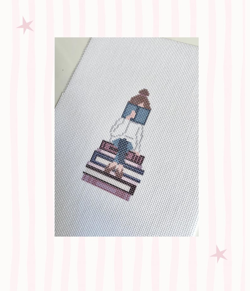 K&ouml;nnte beinhalten: Kreuzstich-Kunstwerk, das eine Person zeigt, die auf einem Stapel B&uuml;cher sitzt und liest. Die Figur ist in einem minimalistischen Stil mit einer Palette aus Blau-, Lila- und Braunt&ouml;nen dargestellt. Der Hintergrund ist eine wei&szlig;e Leinwand mit einem rosa-wei&szlig; gestreiften Rand.