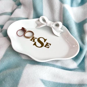 May include: White ceramic ring dish with a white bow on top. The dish has the letters "KSE" in gold lettering on the front.