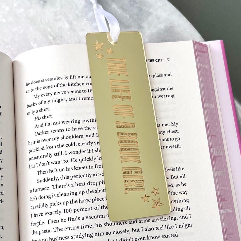 May include: A gold-colored metal bookmark with the text "THE LIFE OF A BOOK GIRL" vertically printed. The bookmark has three small star designs and a white ribbon attached at the top. The bookmark is placed in an open book.