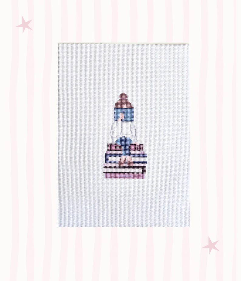May include: Cross-stitch art of a person reading, seated atop a stack of books. The person is wearing a white top and blue jeans. The books are in shades of purple and blue. The background is white with pink stripes and stars.