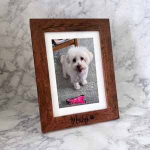 May include: A rectangular wooden picture frame with a dark brown, textured finish. The frame features a white mat and is engraved at the bottom with the name Maisy followed by a paw print icon. Inside the frame is a photograph of a small white dog.