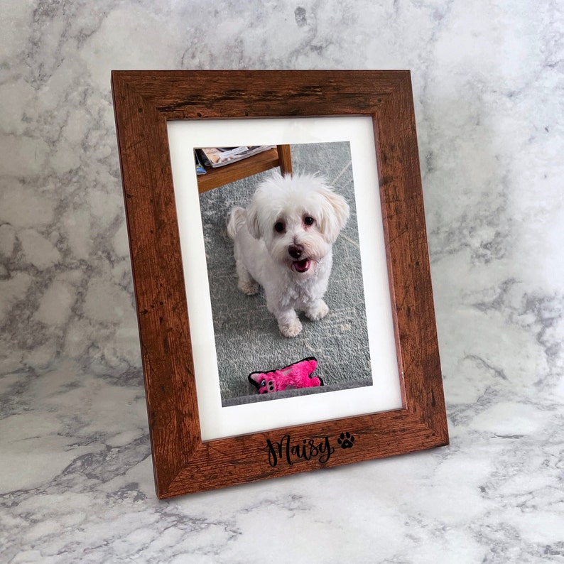 May include: A rectangular wooden picture frame with a dark brown, textured finish. The frame features a white mat and is engraved at the bottom with the name Maisy followed by a paw print icon. Inside the frame is a photograph of a small white dog.