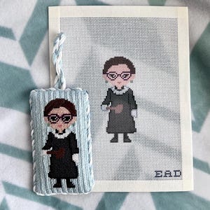 May include: A rectangular needlepoint ornament with a light blue braided cord featuring a cross-stitched figure in a black robe and glasses holding a gavel. Beside it is a matching needlepoint canvas showing the same figure and the letters EAD.