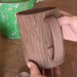 May include: A brown ceramic mug shaped like a tree trunk with a handle. The mug is being held by a hand.