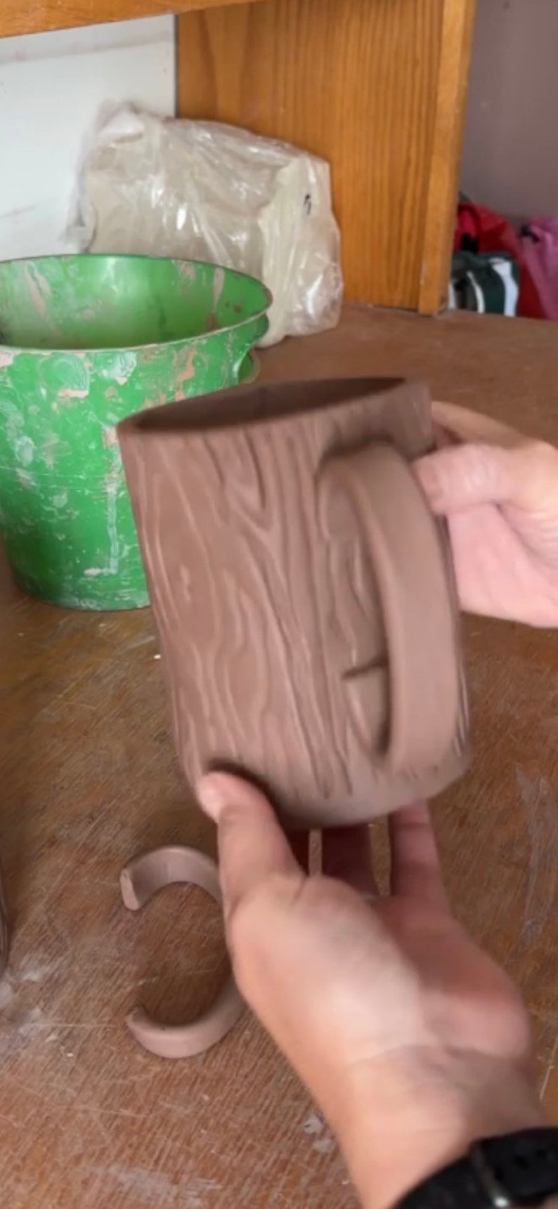 May include: A brown ceramic mug shaped like a tree trunk with a handle. The mug is being held by a hand.