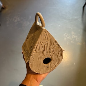 May include: A brown clay birdhouse with a woodgrain texture and a small hole for the birds to enter. The birdhouse has a handle on the top.