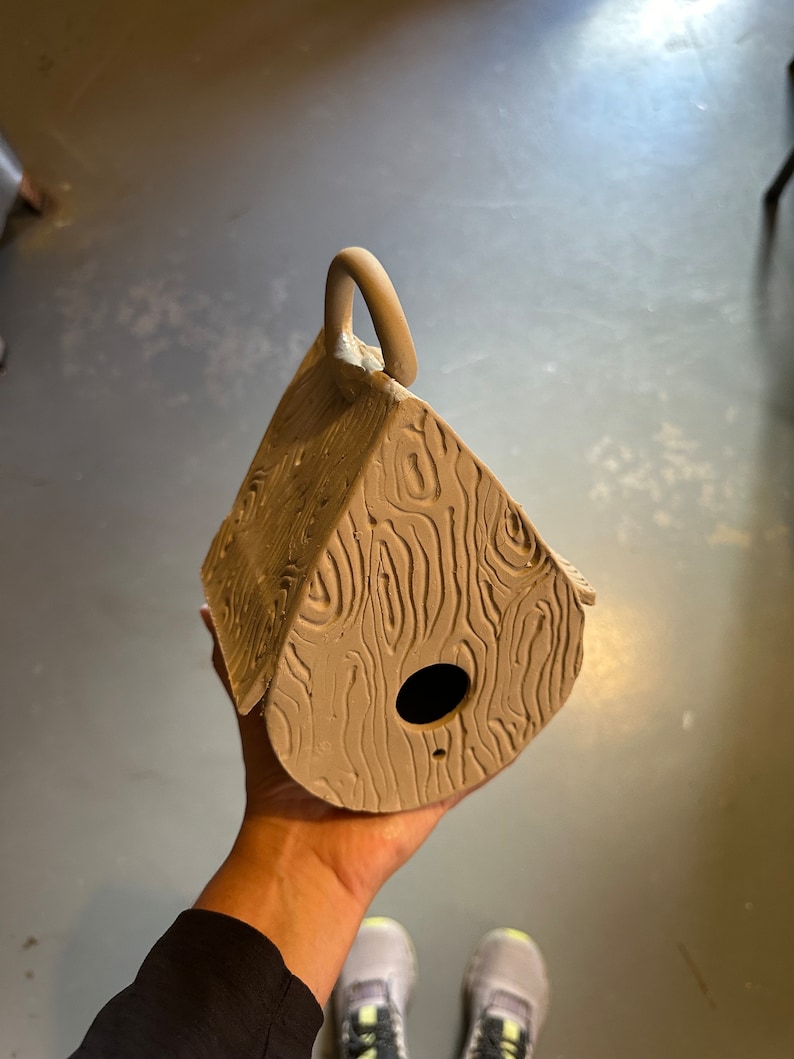 May include: A brown clay birdhouse with a woodgrain texture and a small hole for the birds to enter. The birdhouse has a handle on the top.