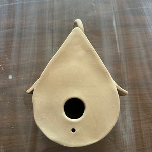 May include: A beige ceramic birdhouse with a round opening and a small hole on the bottom. The birdhouse is shaped like a teardrop and has a handle on the top.
