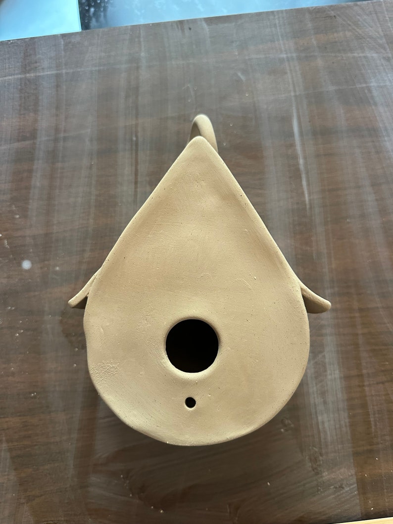 May include: A beige ceramic birdhouse with a round opening and a small hole on the bottom. The birdhouse is shaped like a teardrop and has a handle on the top.