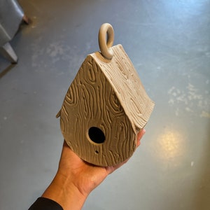 May include: A small, brown, clay birdhouse with a woodgrain texture and a loop on top. The birdhouse has a small hole for birds to enter.