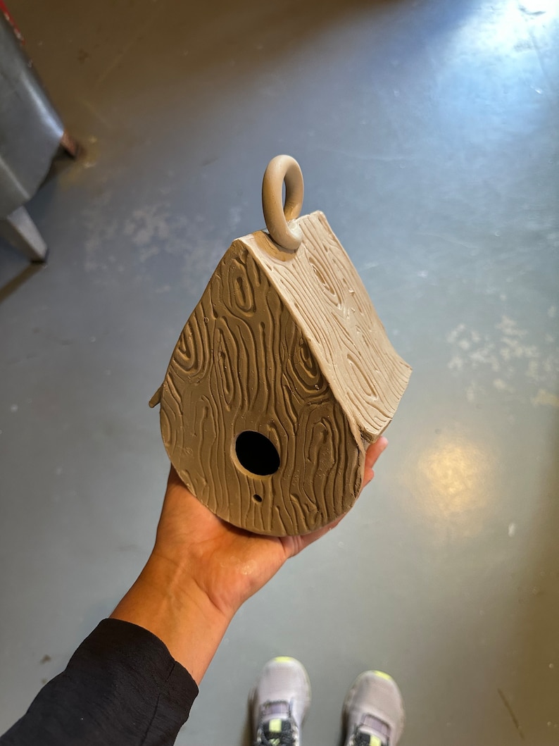 May include: A small, brown, clay birdhouse with a woodgrain texture and a loop on top. The birdhouse has a small hole for birds to enter.