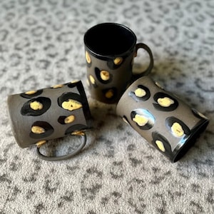 May include: Three ceramic mugs with a black and yellow leopard print design. The mugs are on a gray and white patterned surface.