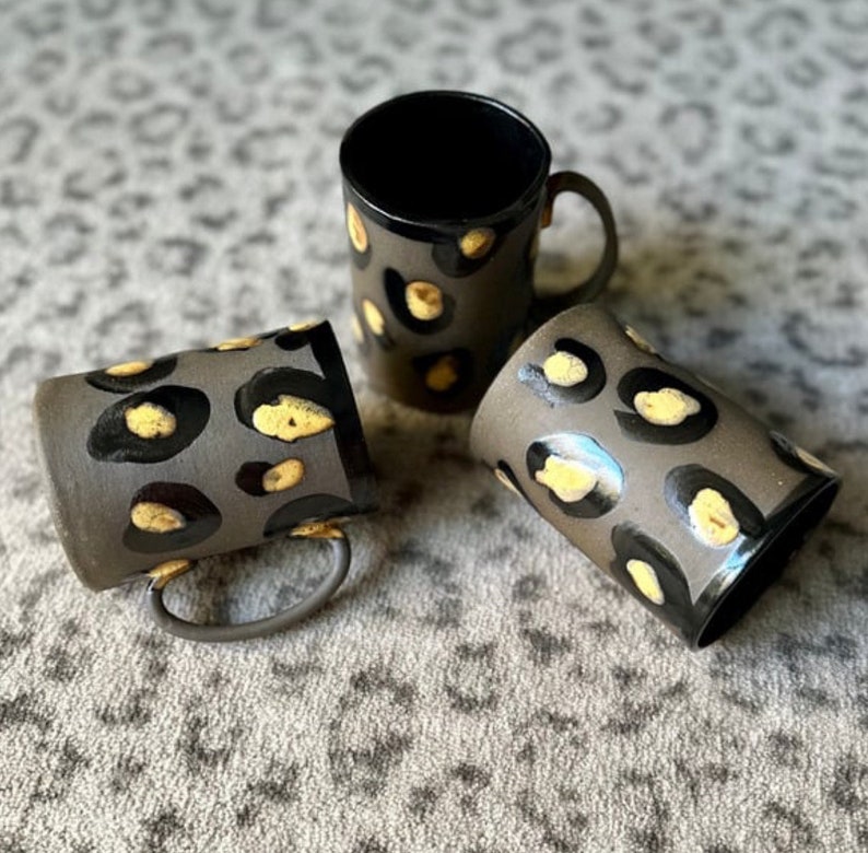 May include: Three ceramic mugs with a black and yellow leopard print design. The mugs are on a gray and white patterned surface.
