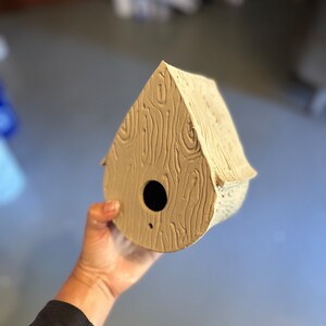 May include: A light brown ceramic birdhouse shaped like a teardrop with a round opening and a woodgrain texture.
