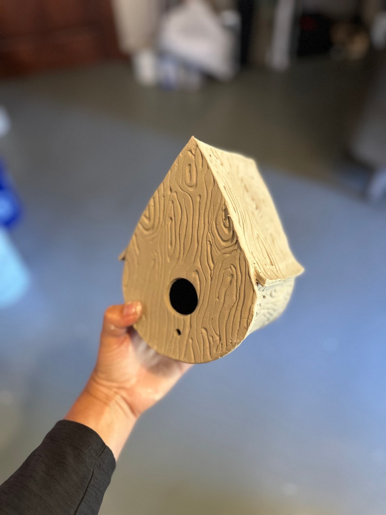 May include: A light brown ceramic birdhouse shaped like a teardrop with a round opening and a woodgrain texture.