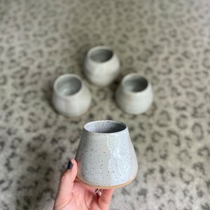 May include: Four white ceramic vases with a speckled glaze. The vases are all the same shape, but different sizes. The vases are on a white and gray patterned rug.
