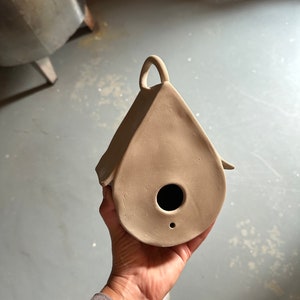 May include: A light brown clay birdhouse with a small hole for the birds to enter. The birdhouse has a handle on the top and is shaped like a teardrop.