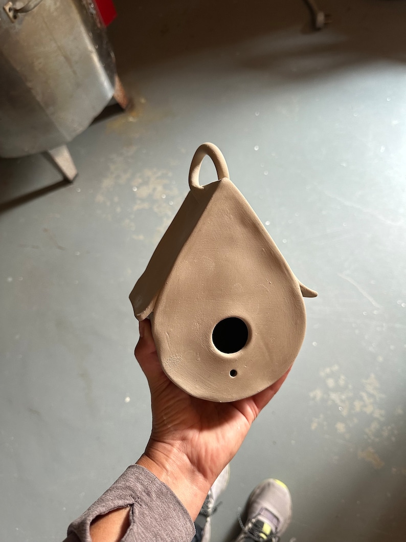May include: A light brown clay birdhouse with a small hole for the birds to enter. The birdhouse has a handle on the top and is shaped like a teardrop.