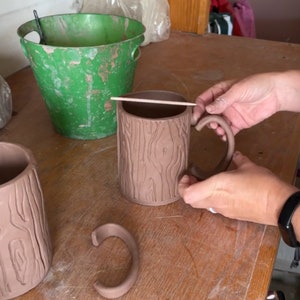 May include: Two brown ceramic mugs with a tree bark texture. One mug has a handle attached, the other has a separate handle. The handle is a simple C shape.