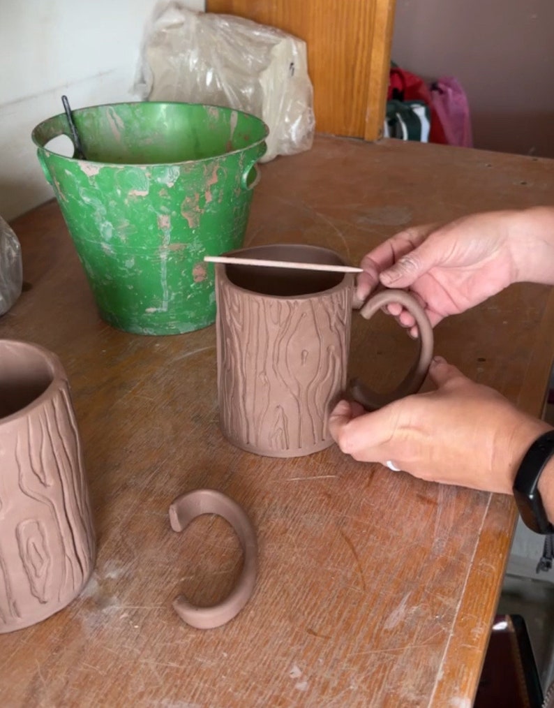 May include: Two brown ceramic mugs with a tree bark texture. One mug has a handle attached, the other has a separate handle. The handle is a simple C shape.