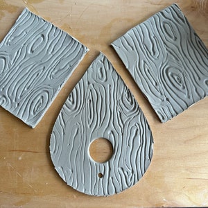 May include: Three pieces of gray clay with a wood grain texture. The clay is shaped into a square, a rectangle, and a teardrop shape with a hole in the center.