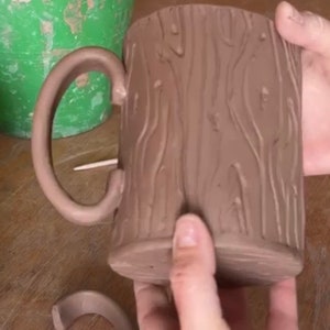 May include: A brown ceramic mug with a tree bark texture. The mug has a handle and is being held by a hand. There is a smaller brown ceramic ring on the table.