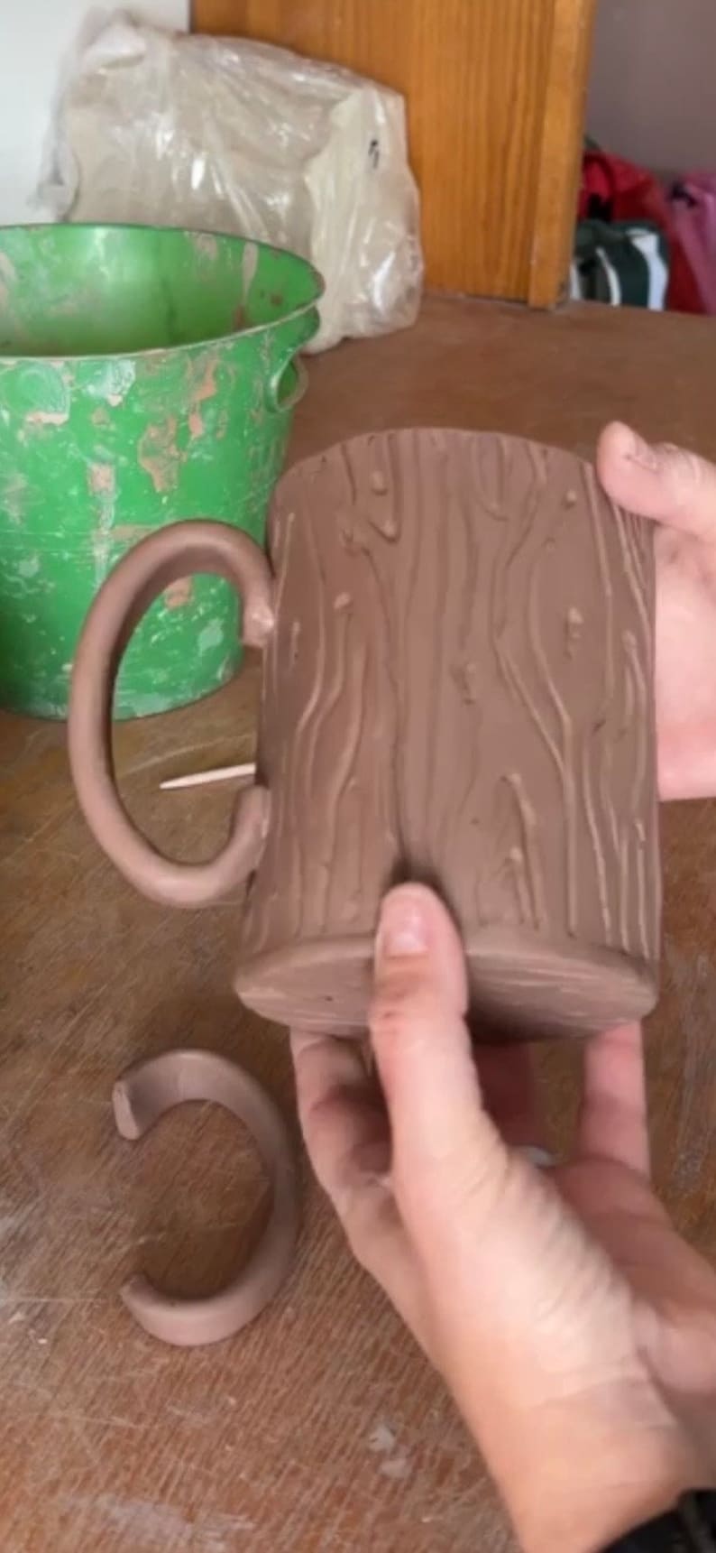 May include: A brown ceramic mug with a tree bark texture. The mug has a handle and is being held by a hand. There is a smaller brown ceramic ring on the table.