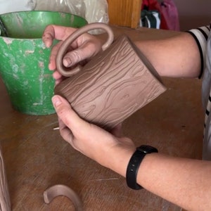 May include: A brown ceramic mug with a wood grain design and a unique handle. The mug is being held by a person's hand.
