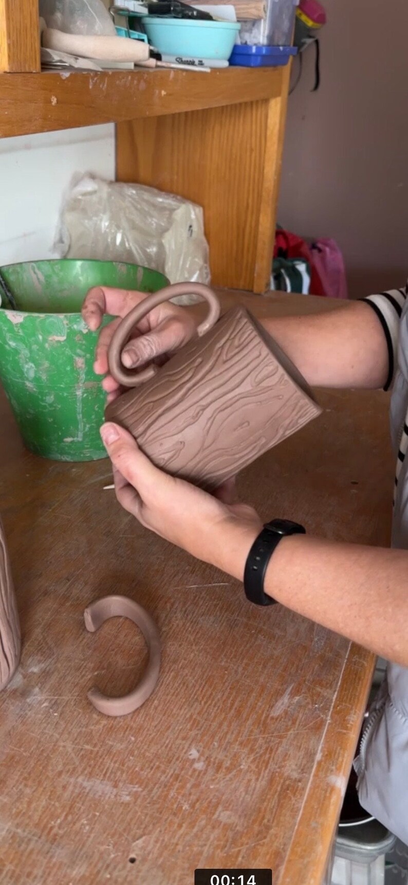 May include: A brown ceramic mug with a wood grain design and a unique handle. The mug is being held by a person's hand.