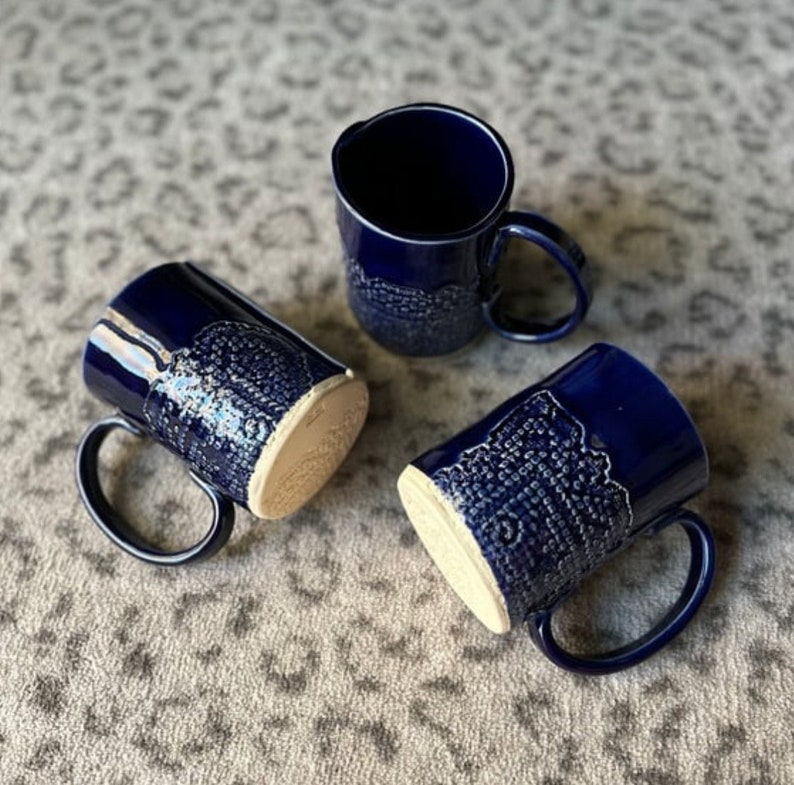 May include: Three blue ceramic mugs with white designs. The mugs have a textured surface and a white rim.