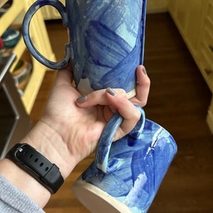 May include: Two blue and white ceramic mugs with abstract brushstroke designs. The mugs are stacked on top of each other.