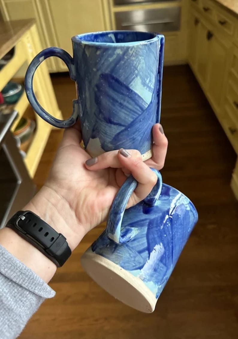 May include: Two blue and white ceramic mugs with abstract brushstroke designs. The mugs are stacked on top of each other.