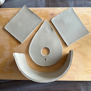 May include: Four pieces of clay pottery on a wooden surface. The pieces are a teardrop shape with a hole in the center, a curved piece, and two square pieces.