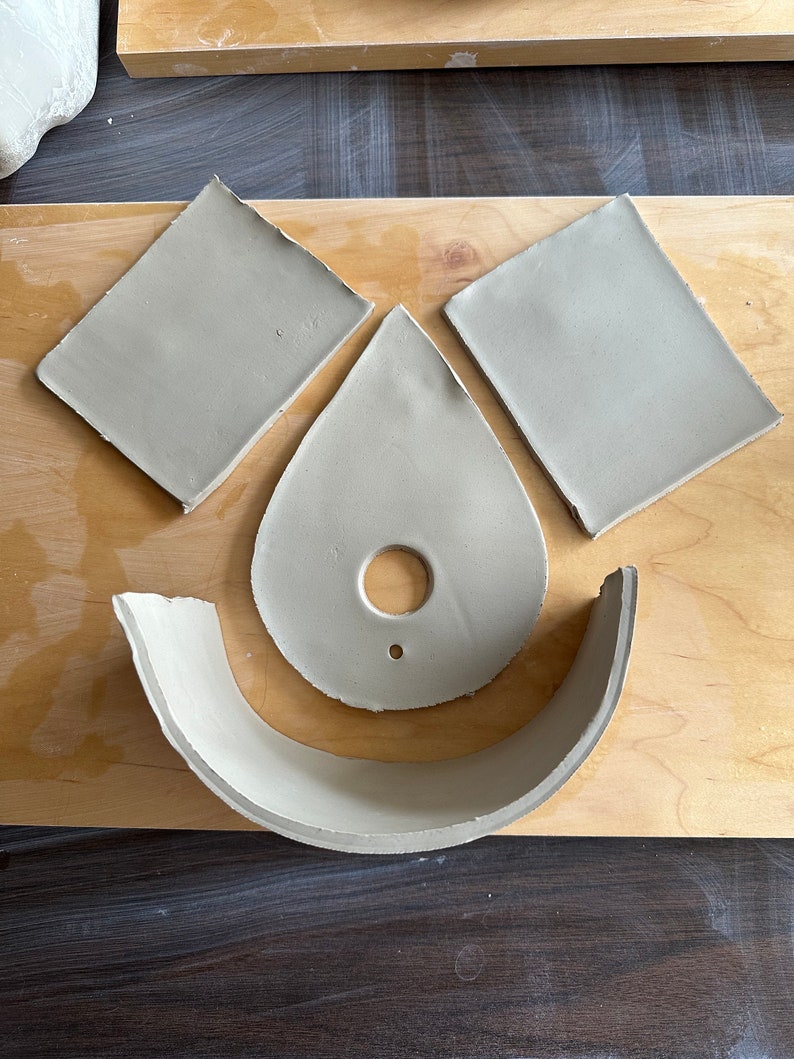 May include: Four pieces of clay pottery on a wooden surface. The pieces are a teardrop shape with a hole in the center, a curved piece, and two square pieces.