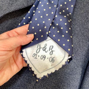 May include: Navy blue tie with a repeating pattern of small yellow polka dots. The tie has a white tag with the initials "J&B" and the date "01.09.06" embroidered on it. The tie is held over a dark blue textured fabric.
