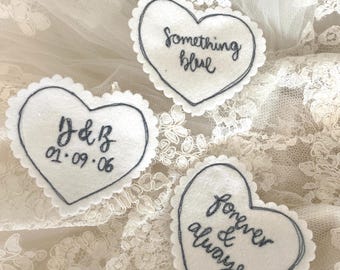 May include: Three white heart-shaped felt patches with dark gray embroidered details. One says "Something blue", another has initials and a date, and the third says "forever & always". The patches have scalloped edges.